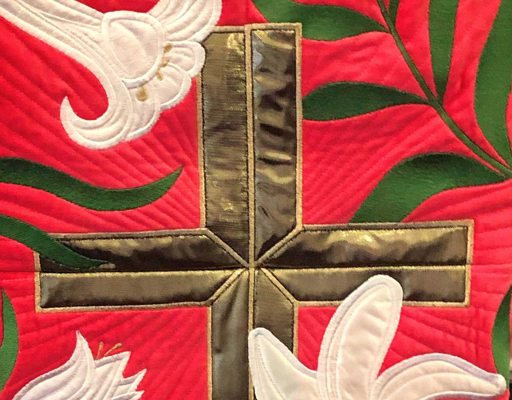 Close-up of a quilted fabric featuring a shiny gold cross, white lilies, and green leaves on a red background.