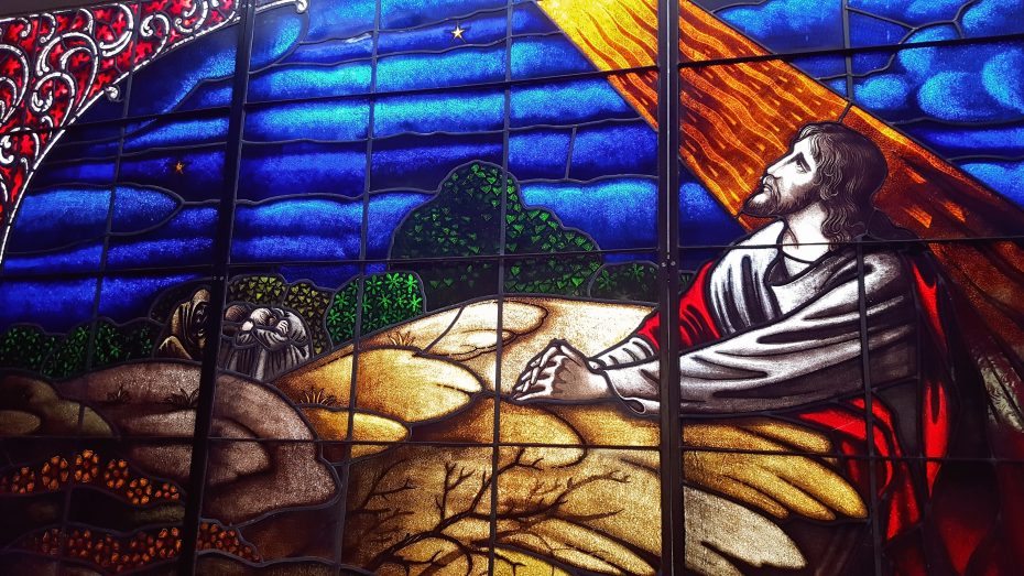 Stained glass window depicting Jesus praying on a rock with trees, a night sky, and beams of light shining down.