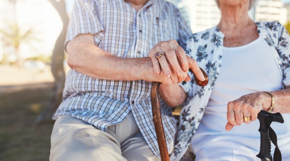 An elderly couple sitting side by side, holding walking sticks. The man's hand rests on top of the woman's hand. Both are dressed in casual clothing.