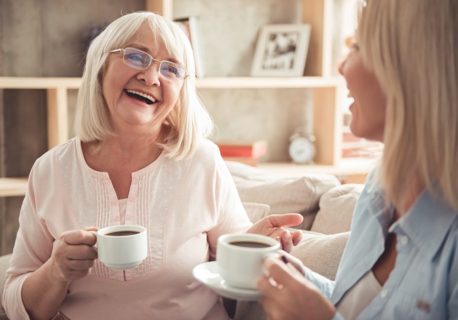 Two women sitting on a sofa, smiling and holding cups of coffee. A bookshelf is in the background.