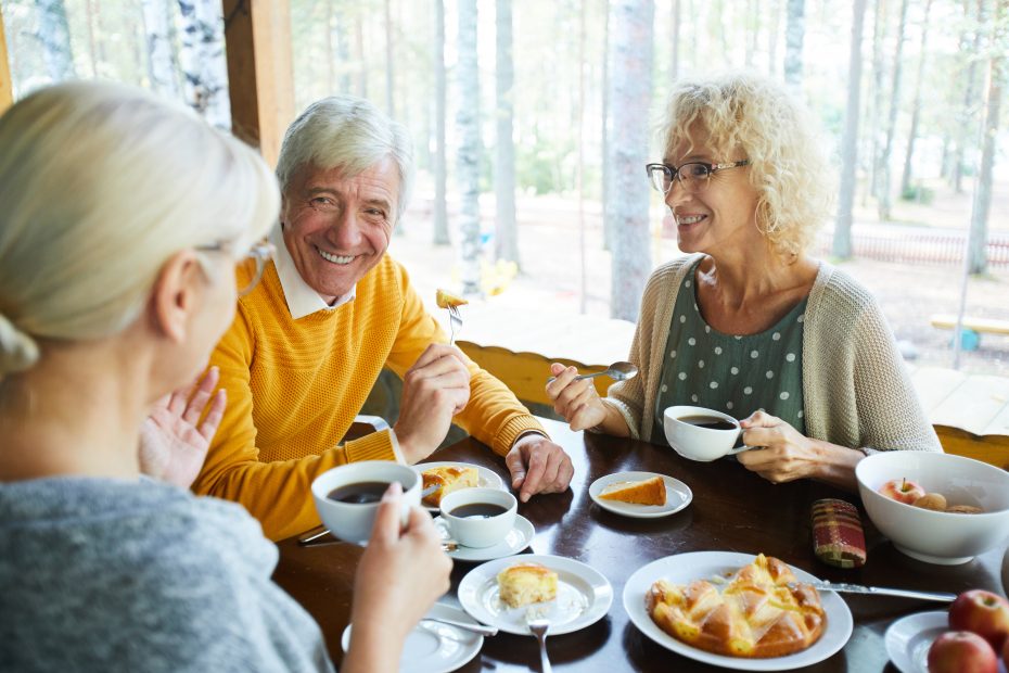 Three people enjoy a meal together at a wooden table outdoors, with coffee, cake, and conversation. Trees are visible in the background.