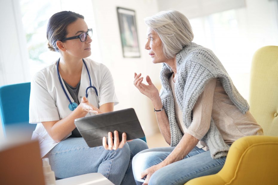 A doctor holding a tablet discusses something with an elderly woman in a cozy setting with natural light.