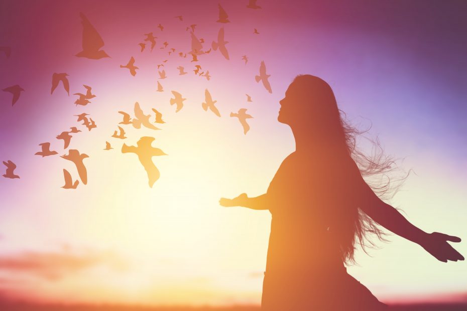 Silhouetted woman with outstretched arms faces a flock of birds at sunset, with the sky transitioning from purple to orange hues.