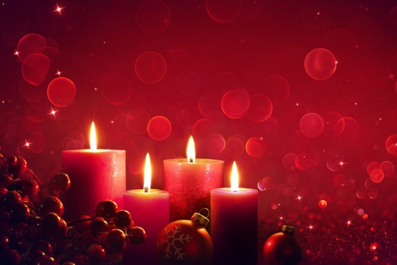 Four lit red candles, with red ornaments and festive lights in the background.