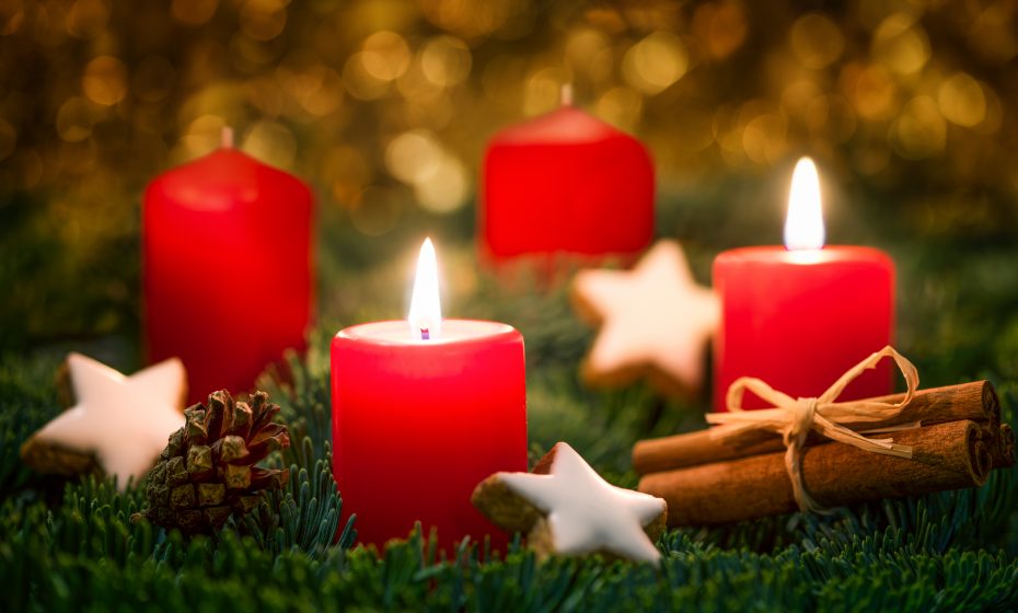 Four red candles, two lit and two unlit, are placed on a bed of greenery surrounded by white star-shaped ornaments, pinecones, and cinnamon sticks, set against a blurry, golden background.