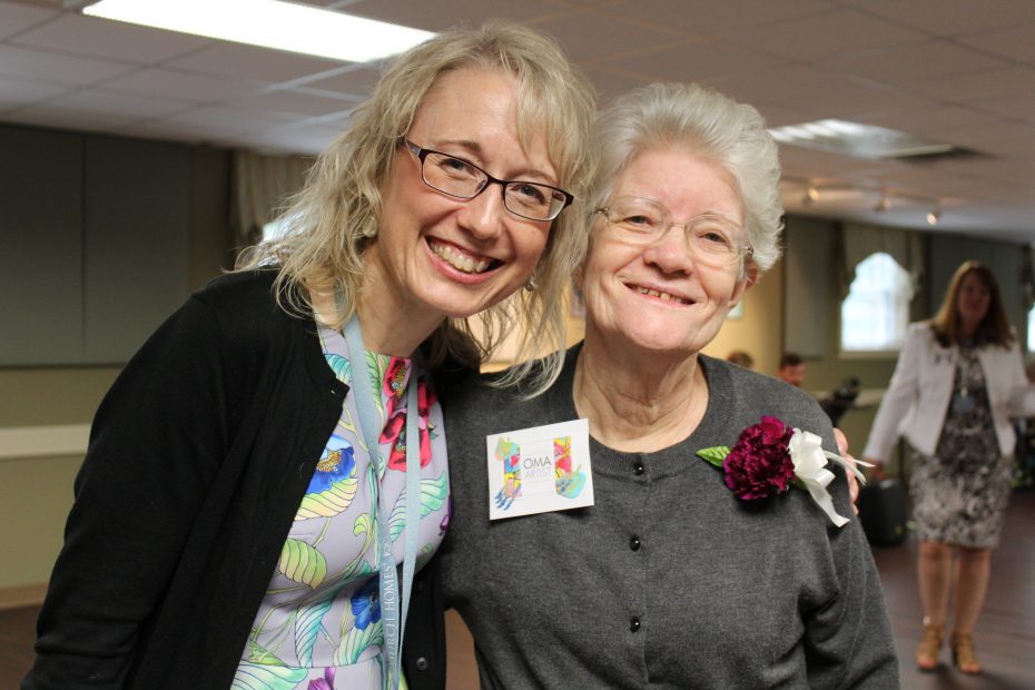 Two women smiling, one older wearing glasses and a flower pin, the other younger with glasses and a name tag. They are standing closely in an indoor setting.
