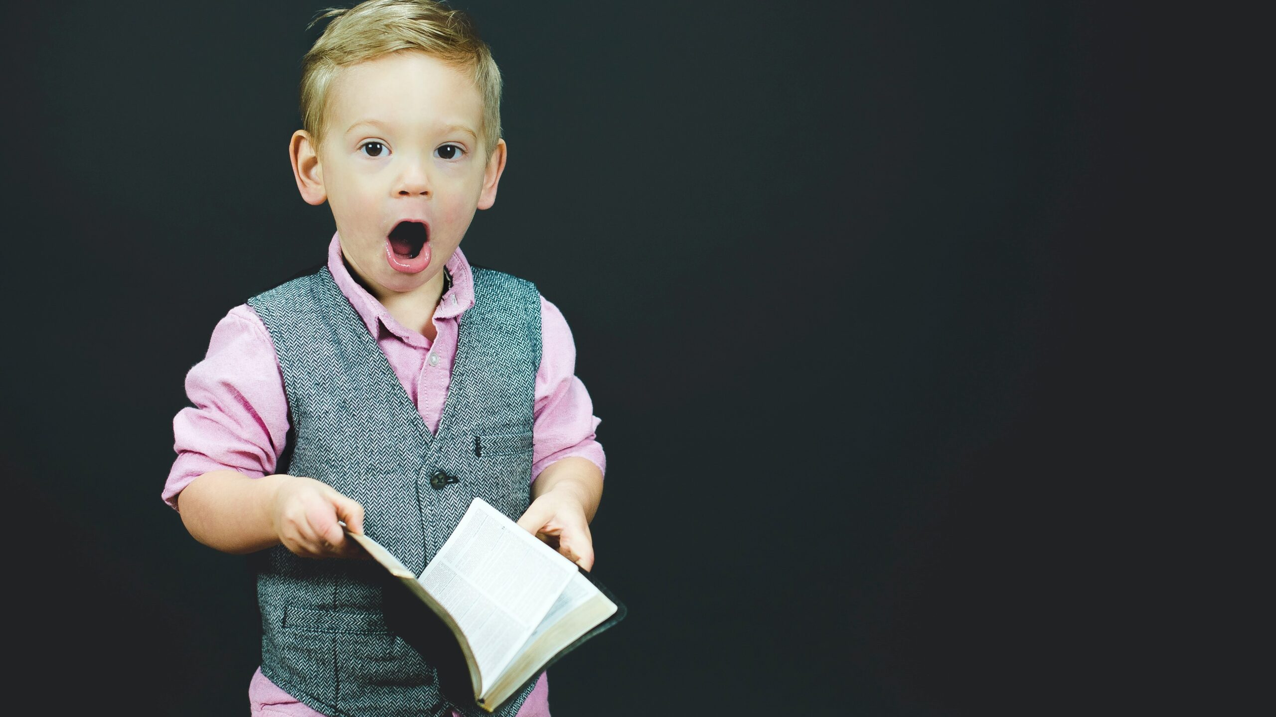 A young boy wearing a pink shirt and grey vest stands against a black background, holding an open book, with a surprised expression on his face.