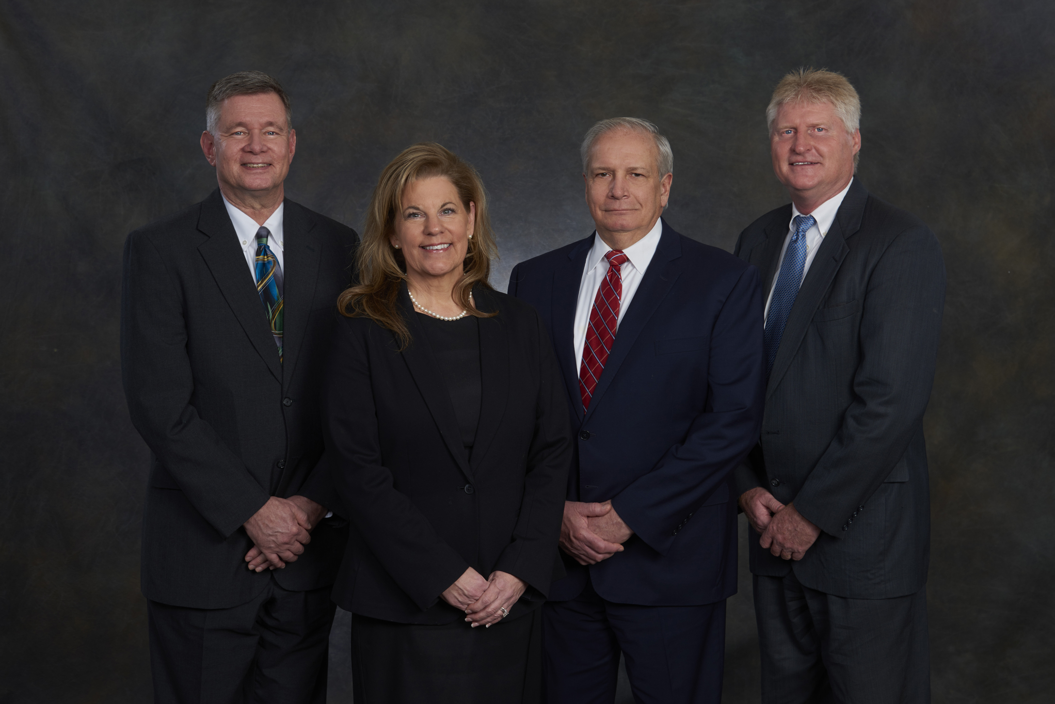 Four individuals in business attire stand side by side against a dark, neutral backdrop for a professional portrait, embodying the spirit of restructures for growth.