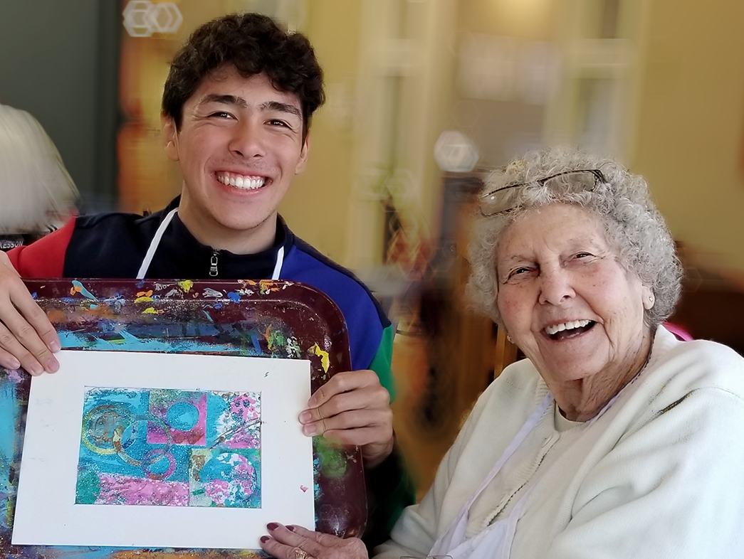 A young man and an elderly woman smile at the camera; the young man holds a colorful piece of art on a tray, showcasing their journey of opening minds through art.