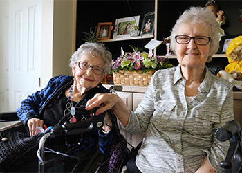 Two elderly women, one in a wheelchair and the other standing beside her, smile at the camera. They are indoors with a shelf of family photos and a basket of flowers in the background.