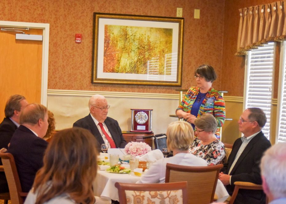 A group of people sit around a table set for a meal. A woman stands and speaks, while an elderly man and others listen attentively. A clock and framed artwork are on the wall in the background.