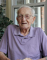An elderly man wearing glasses and a lavender polo shirt sits in front of a window with a slight smile. A plant is partially visible in the background. An elderly man wearing glasses and a lavender polo shirt sits in front of a window with a slight smile. A plant is partially visible in the background.