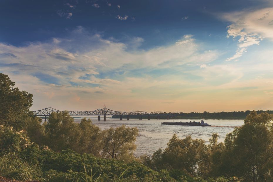 A barge travels along a wide river, passing under a distant bridge framed by a partly cloudy sky and lush greenery on the riverbanks.