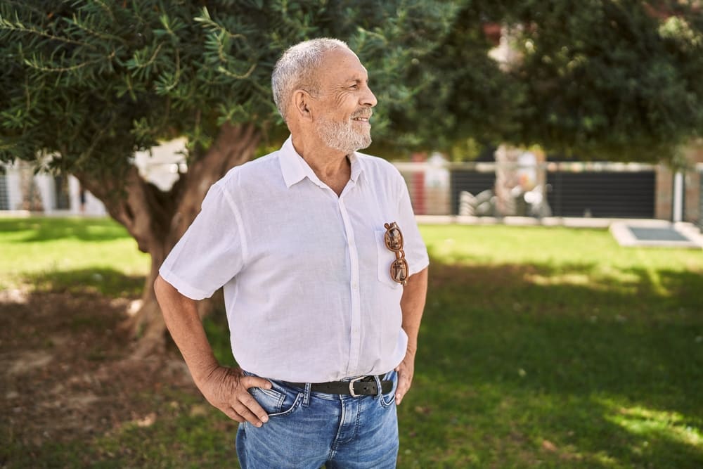 An older man with a beard stands outdoors in a park, wearing a white shirt and blue jeans, holding sunglasses in his left hand like he just stepped out of the internal career center.