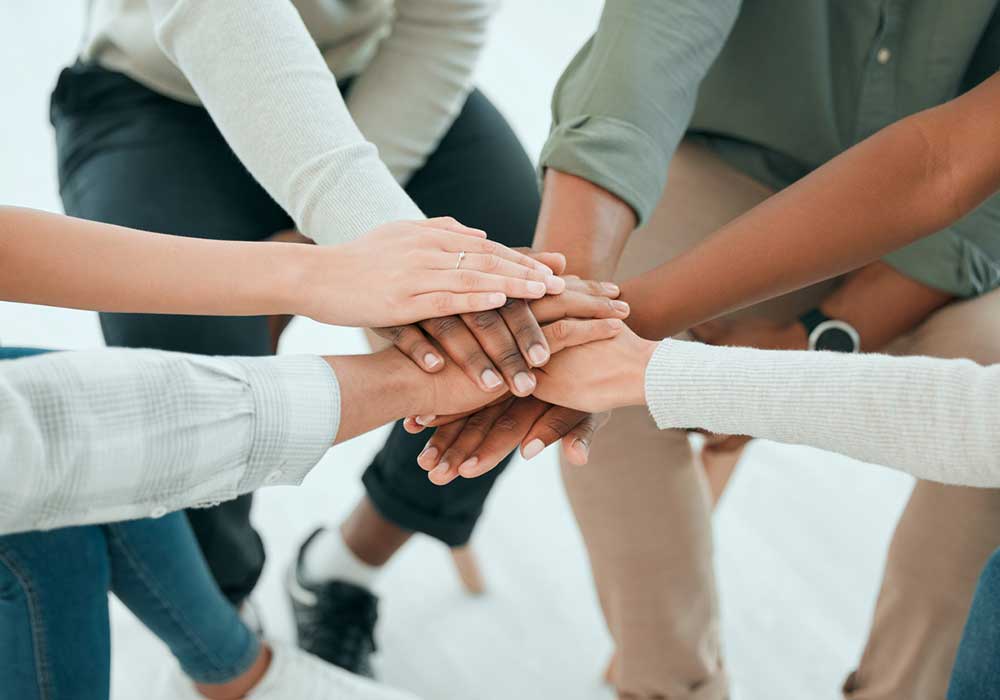A group of people seated in a circle place their hands on top of each other in the center, symbolizing unity and teamwork.
