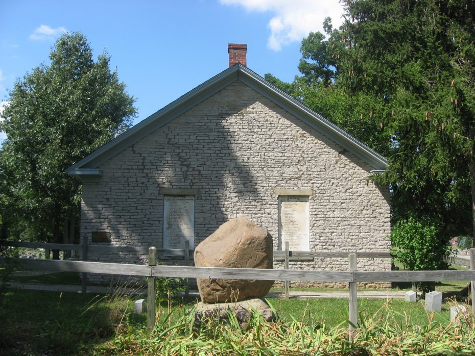 A small stone building with a gabled roof stands behind a wooden fence and a large rock in the foreground, surrounded by trees and greenery.