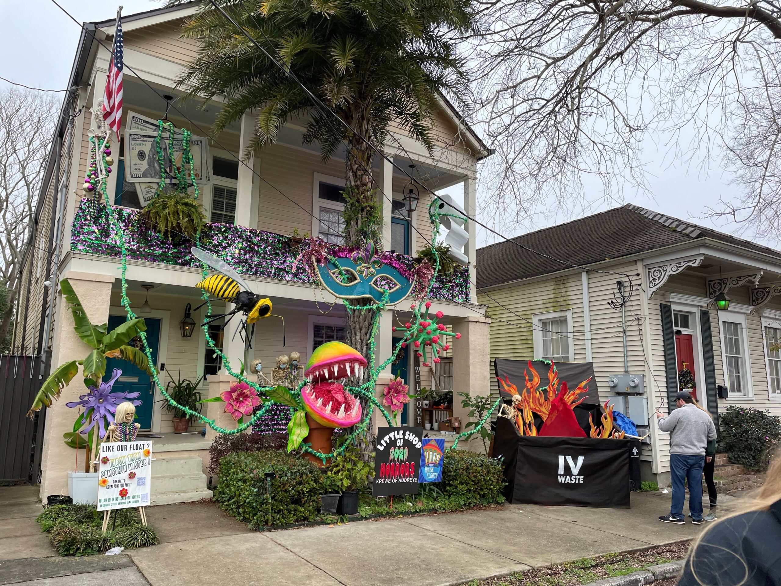 A house decorated with colorful and whimsical plants and flowers for a celebration, with "House Floats" and "IV Waste" signs visible. A person stands by the house on the right, partially out of view.