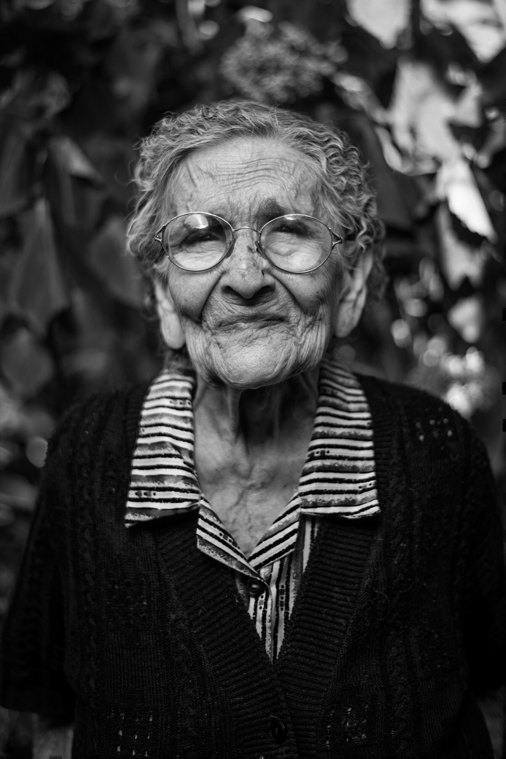 Black and white portrait of an elderly person with glasses and curly hair, wearing a striped shirt and dark cardigan, standing outdoors.