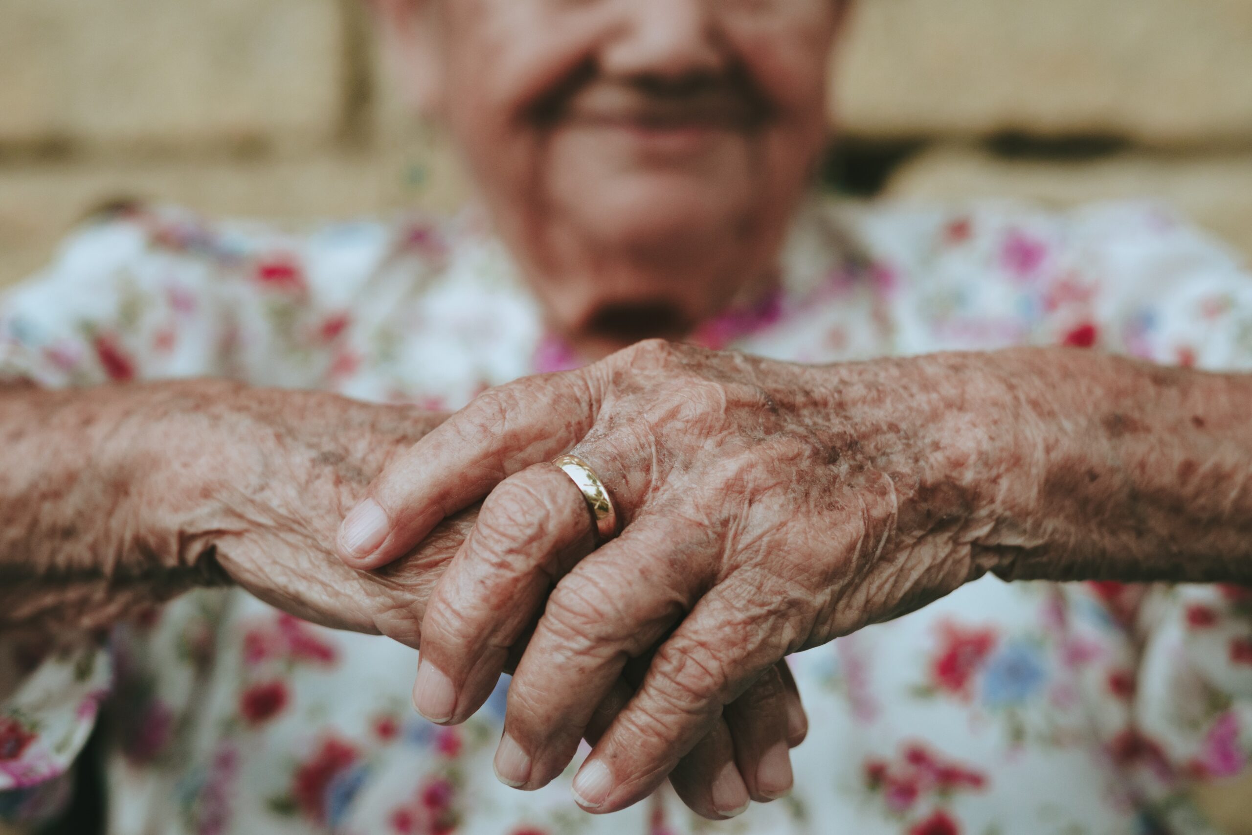 Elderly person with a floral garment showcasing wrinkled hands folded, wearing a gold ring on the left hand, with a blurred background.