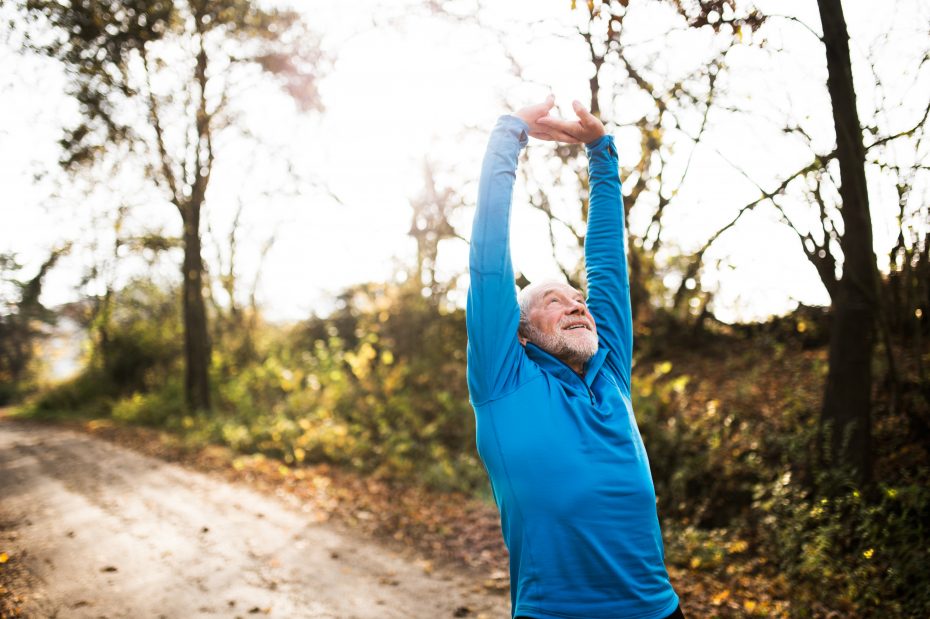 A man in a blue long-sleeve shirt stretches with arms overhead on a wooded path on a sunny day.