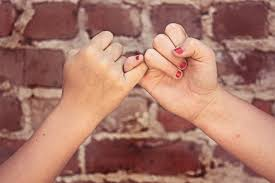 Two hands with interlocked pinkies in front of a red brick wall background. Both have painted nails.