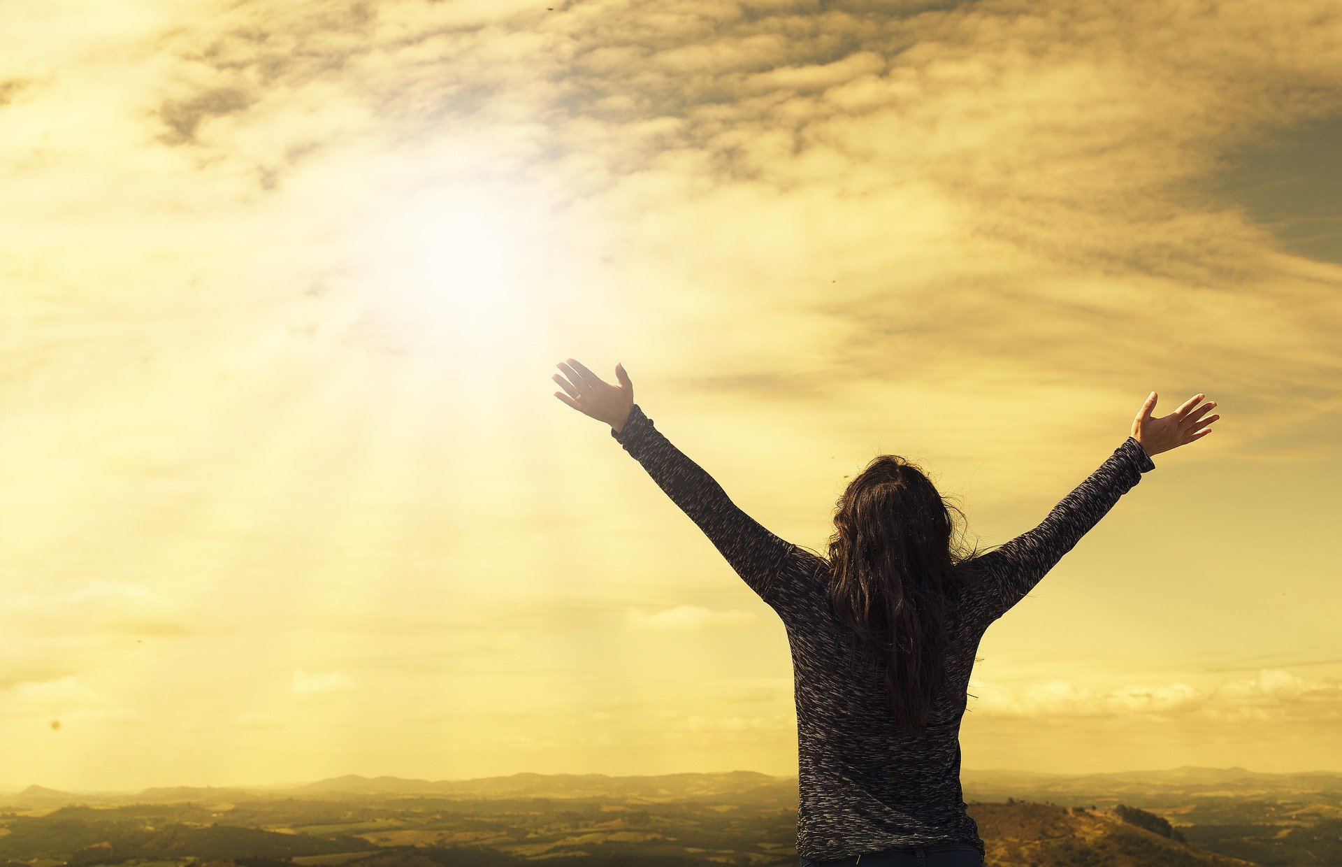 A person stands with arms raised toward the sky, facing a bright sun and an expansive, cloudy landscape.